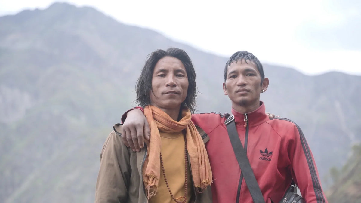 Two Nepali honey hunters arm-in-arm on a mountain trail, ropes and harvest gear on their backs